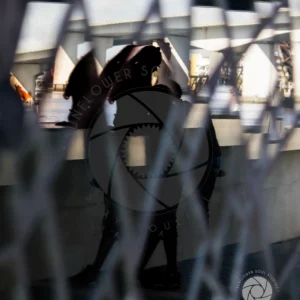The Couple 2, Two silhouetted figures standing together in quiet companionship, viewed through a chain-link fence against an industrial backdrop along the St. Johns River, shot in muted natural light.